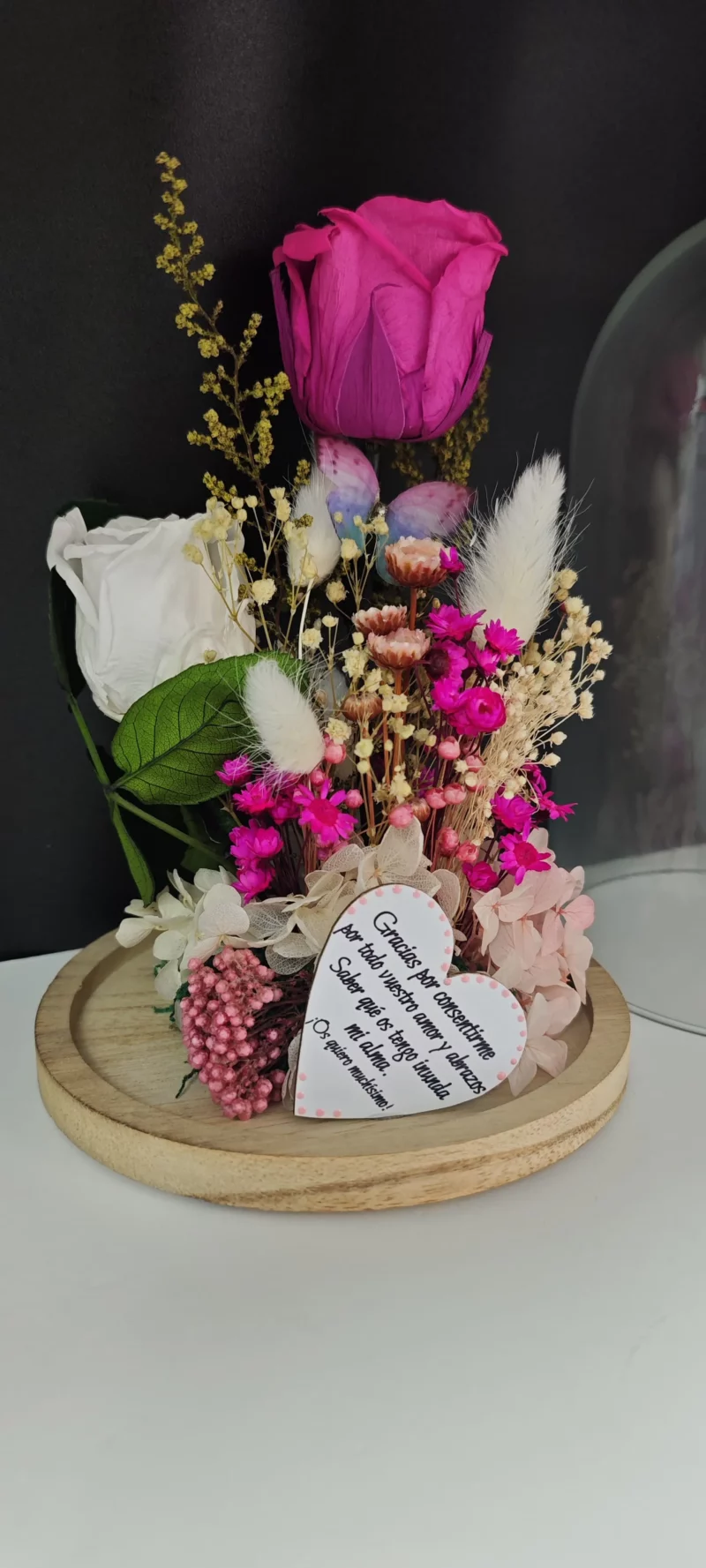 Cúpula de cristal con rosas preservadas en blanco y fucsia, flores secas y corazón de madera grabado.