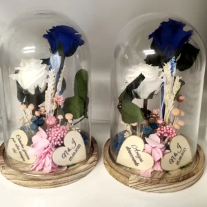 Cúpula de cristal con rosas preservadas en tonos azul y blanco, decorada con corazones grabados y base de madera.