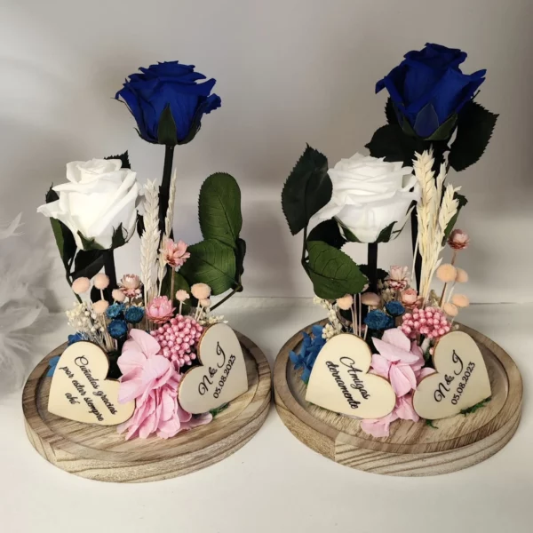 Cúpula de cristal con rosas preservadas en tonos azul y blanco, decorada con corazones grabados y base de madera.