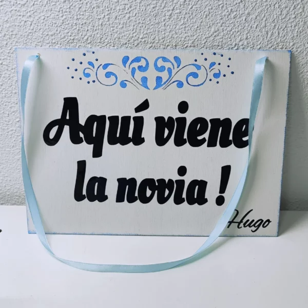 Cartel de madera pintado en blanco con texto “Aquí viene la novia” en negro y detalles azules, con cinta azul para colgar
