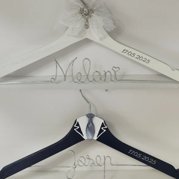 Pareja de perchas novios elegantes: novia blanca con encaje y broche de cristal; novio azul marino con cuello blanco y corbata gris, nombres en alambre