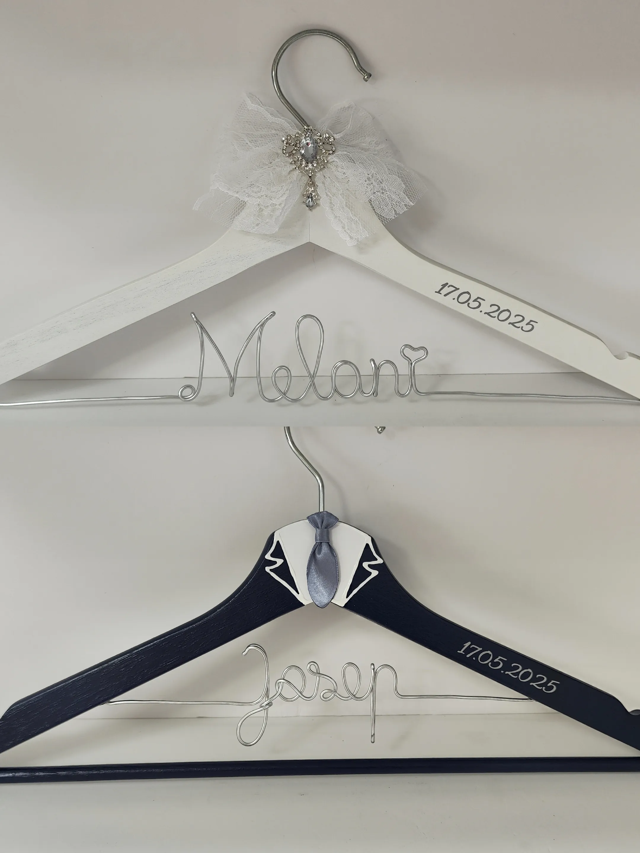 Pareja de perchas novios elegantes: novia blanca con encaje y broche de cristal; novio azul marino con cuello blanco y corbata gris, nombres en alambre