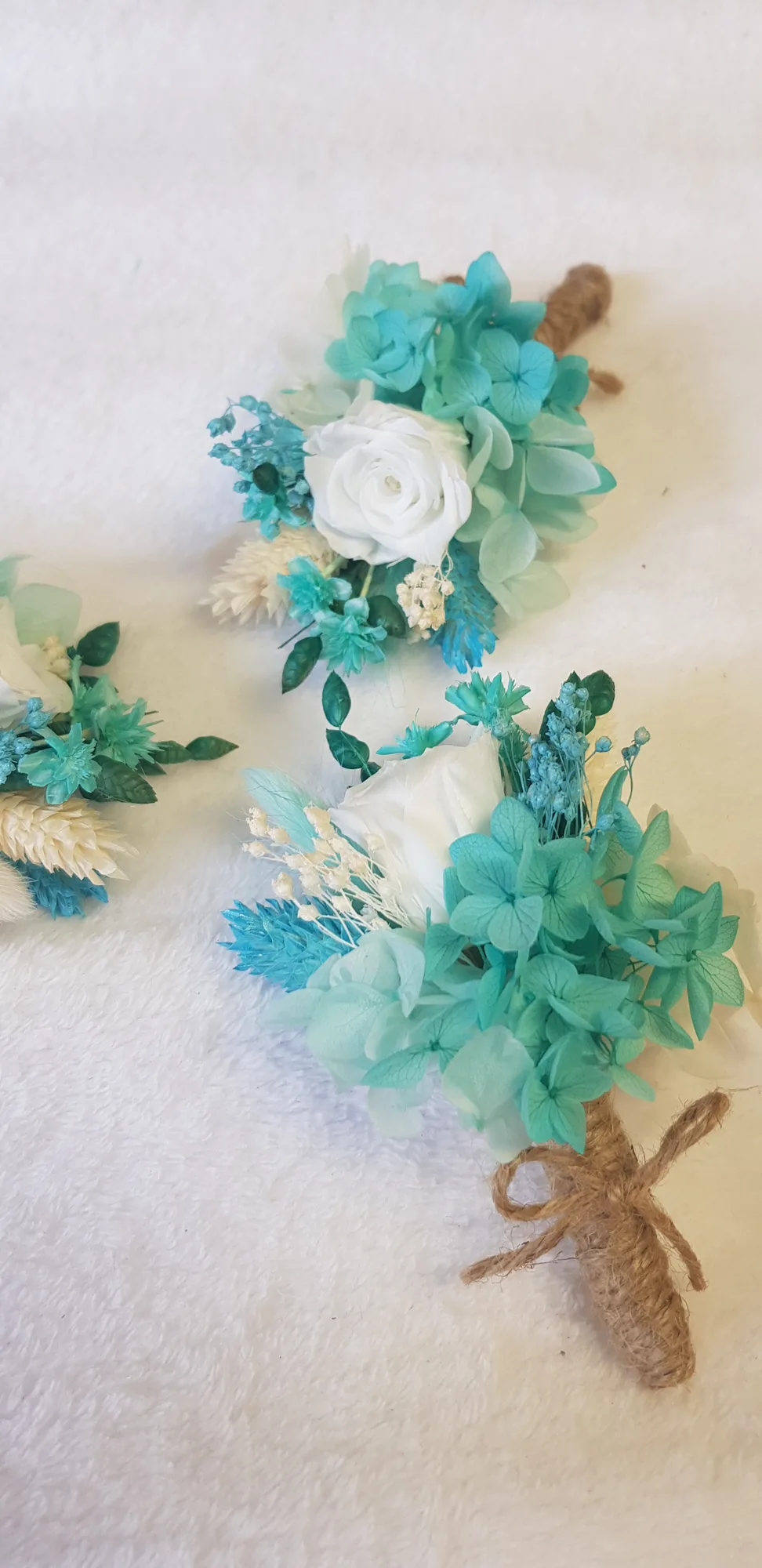 Varias unidades del prendido de novio de flores preservadas turquesa y blanco, con tallo envuelto en yute